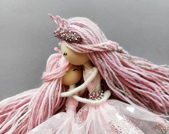 May include: Two handmade dolls, embracing, with pink yarn hair and dresses. The dolls have wooden heads and are adorned with sparkly embellishments. One doll wears a crown. The dresses are made of tulle and have silver accents.