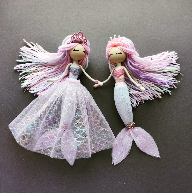 May include: Two handmade dolls dressed as mermaids. One doll wears a pink and purple dress with a crown and the other doll wears a pink and blue mermaid tail with a crown.