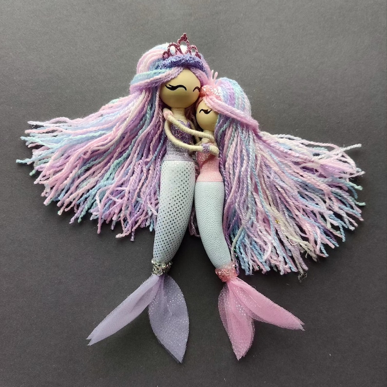 May include: Two handmade mermaid dolls with long, colorful yarn hair. The dolls are wearing blue and pink fishtail tails with tulle accents. The dolls are hugging each other.
