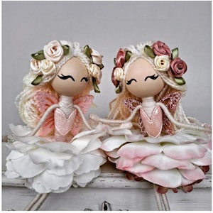 Könnte beinhalten: Zwei handgefertigte Feenpuppen mit Wollhaaren, Blumenkränzen und rosa-weißen Blumenröcken. Die Puppen haben bemalte Gesichter und Glitzerflügel.