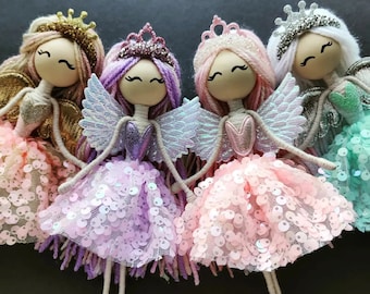 May include: Four handmade fairy dolls with yarn hair, sequined skirts, and glittery wings. Each doll has a wooden head, a crown, and a smiling face. The dolls are in shades of pink, purple, gold, and green.