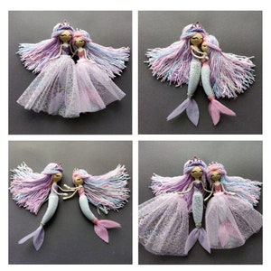 May include: Two handmade mermaid dolls with long, colorful yarn hair, wearing pink and purple tulle skirts and crowns. The dolls are holding hands and have painted faces.
