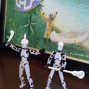 May include: Two articulated white and black figures, each holding a lacrosse stick, stand before a framed painting depicting a Native American scene. The painting shows a canoe, a figure with arms raised, and a colourful woven design.