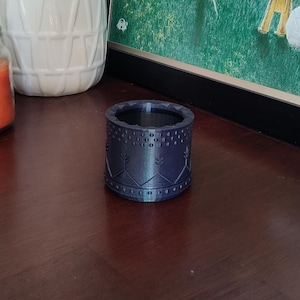 May include: A dark blue cylindrical container with a detailed pattern of dots and stylized tree-like shapes. The container is made of a solid material and sits on a dark wooden surface. The design suggests a decorative or functional item.
