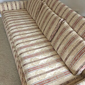 May include: A striped beige sofa with a floral pattern and four matching pillows. The sofa has a tufted back and a rolled armrest.