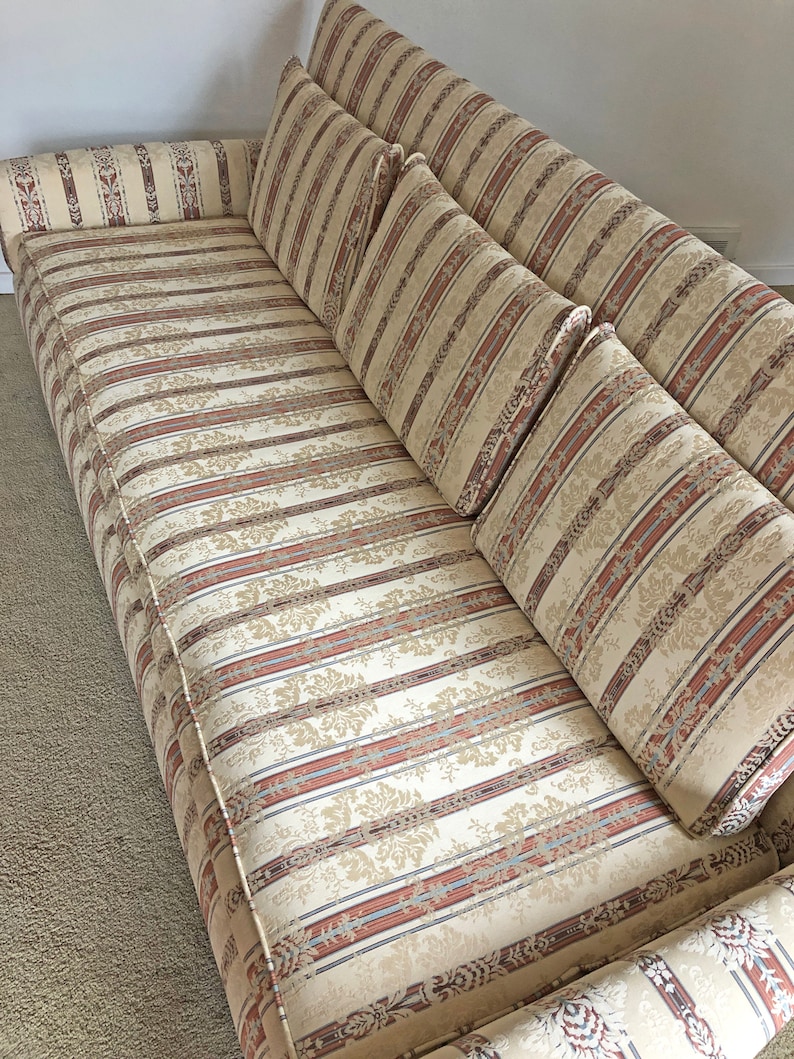May include: A striped beige sofa with a floral pattern and four matching pillows. The sofa has a tufted back and a rolled armrest.