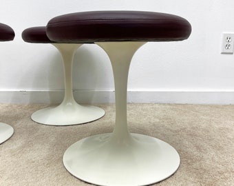 May include: Three mid-century modern stools. Each stool has a dark brown, oval-shaped seat and a white, tulip-shaped base. The stools are placed on a light-colored carpet against a white wall.