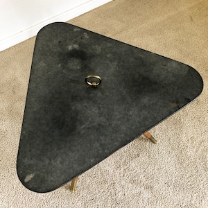 May include: A black triangular table with a brass ring handle on the top. The table has three legs and is sitting on a beige carpet.