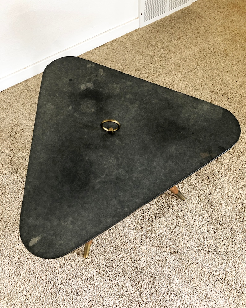 May include: A black triangular table with a brass ring handle on the top. The table has three legs and is sitting on a beige carpet.