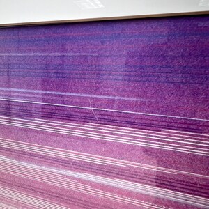 May include: Abstract art print with a gradient of purple and pink hues. Horizontal white lines create a sense of movement across the textured surface. The artwork is framed with a thin, light-colored border.