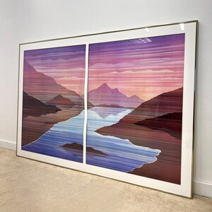 May include: Large framed artwork featuring a landscape scene. The artwork depicts mountains, a lake, and a sunset with hues of purple, pink, and blue. The artwork is encased in a gold-colored frame and is displayed on a wall.
