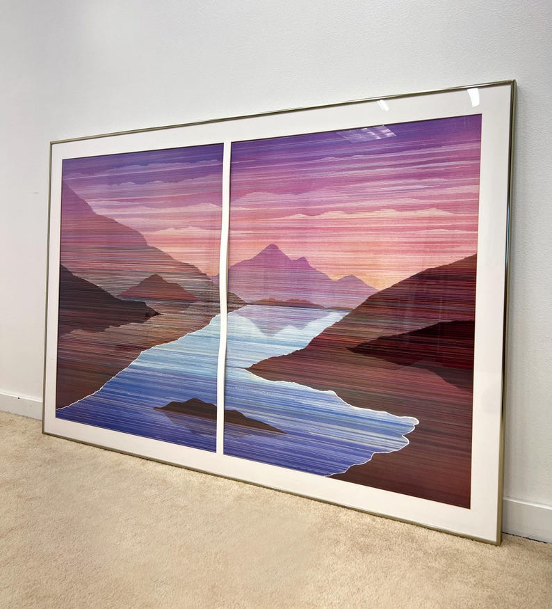 May include: Large framed artwork featuring a landscape scene. The artwork depicts mountains, a lake, and a sunset with hues of purple, pink, and blue. The artwork is encased in a gold-colored frame and is displayed on a wall.