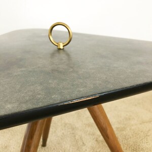 May include: A gray, square-shaped side table with a black edge and a brass ring handle on top. The table has four wooden legs.