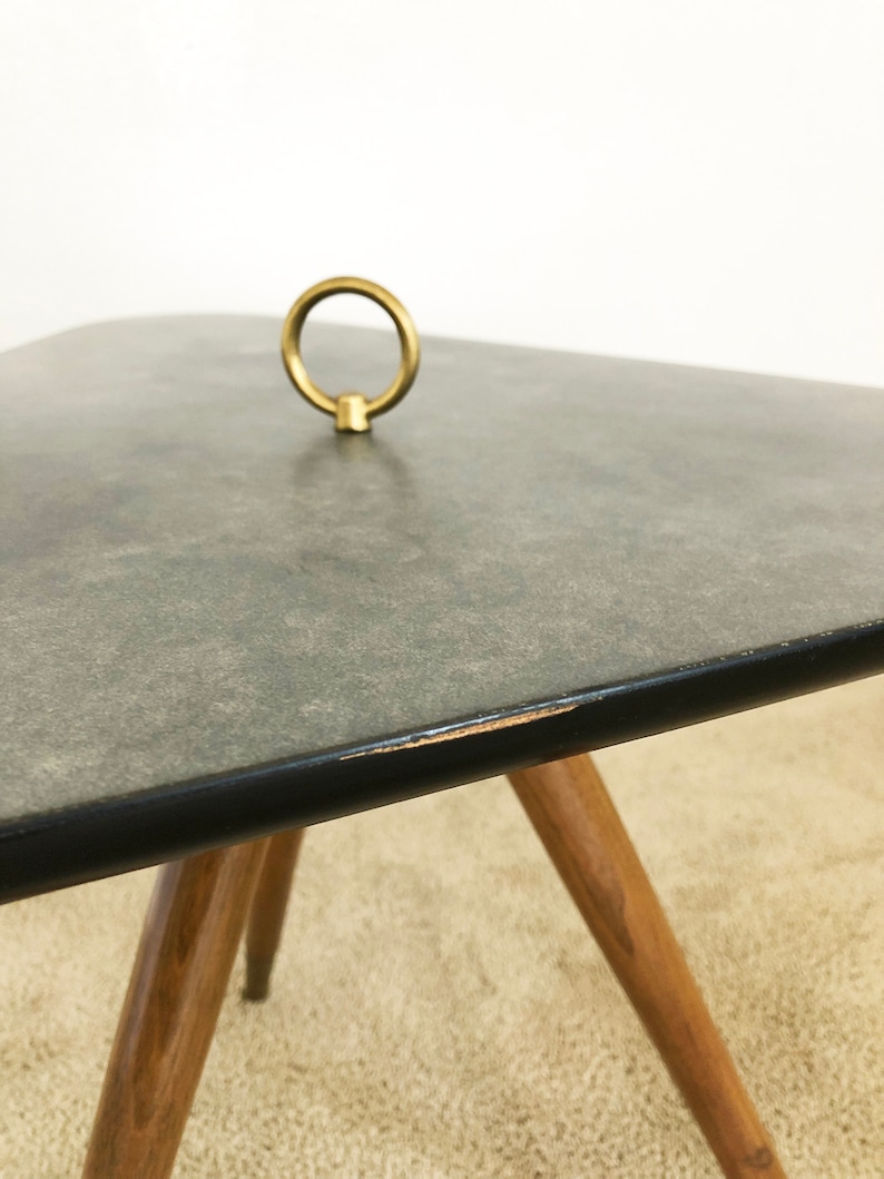 May include: A gray, square-shaped side table with a black edge and a brass ring handle on top. The table has four wooden legs.
