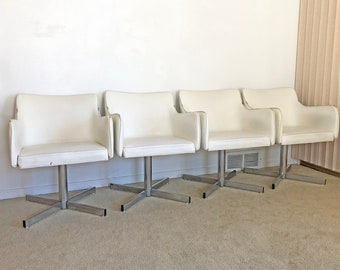 May include: Four white swivel chairs with chrome bases. The chairs are arranged in a row on a light beige carpet.