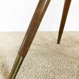May include: Close-up of the legs of a wooden tripod table with brass feet. The table legs are made of dark wood and have a smooth finish. The brass feet are round and have a polished finish.