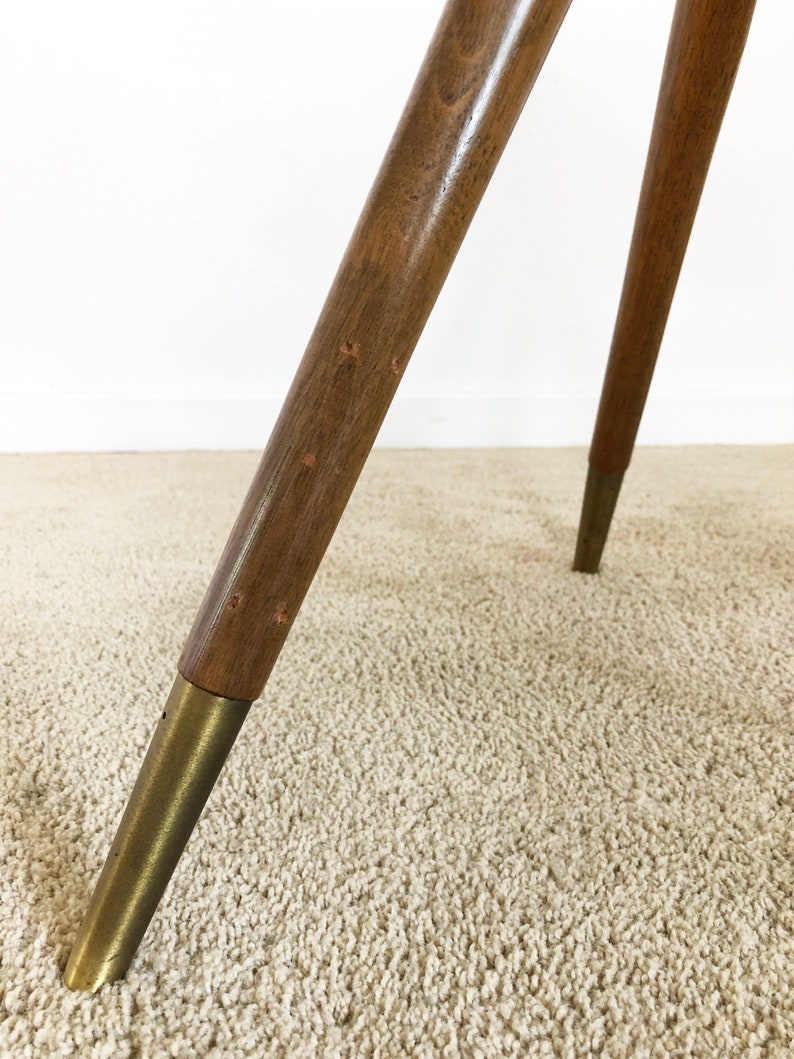 May include: Close-up of the legs of a wooden tripod table with brass feet. The table legs are made of dark wood and have a smooth finish. The brass feet are round and have a polished finish.