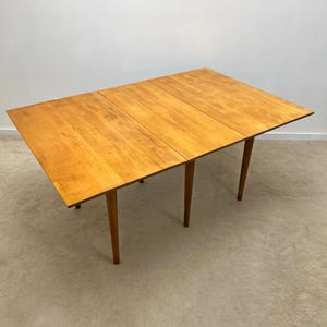 May include: A light brown, mid-century modern drop-leaf table. The table has a rectangular top with three sections and four tapered legs. The table is made of wood and has a natural finish. The drop-leaf is extended on one side.