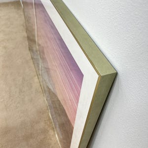 May include: Large framed artwork featuring a landscape scene with a gradient of pink, purple, and white hues. The frame is gold-colored, and the artwork is displayed against a white wall.