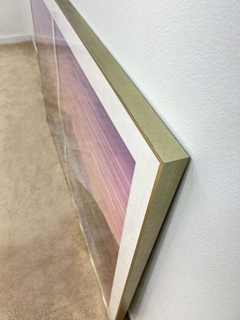 May include: Large framed artwork featuring a landscape scene with a gradient of pink, purple, and white hues. The frame is gold-colored, and the artwork is displayed against a white wall.
