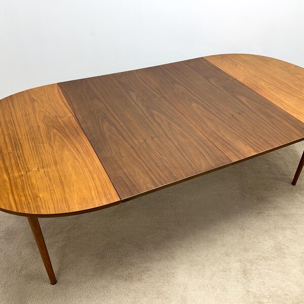 May include: Oval wooden dining table with a two-tone finish. The tabletop is a combination of light and dark brown wood, supported by four slender, angled legs. The table is approximately 183 cm long.