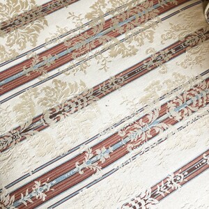 May include: Close-up of a cream-colored fabric with a striped pattern. The stripes are brown, blue, and white. The fabric has a subtle floral design.