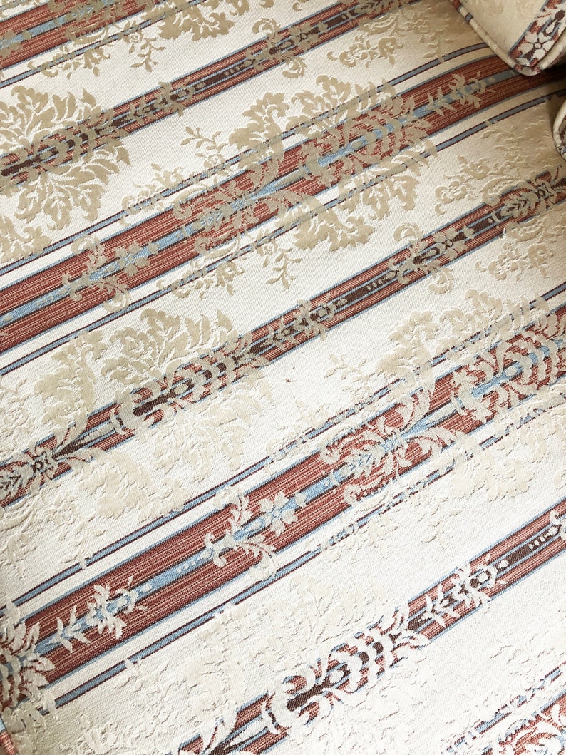 May include: Close-up of a cream-colored fabric with a striped pattern. The stripes are brown, blue, and white. The fabric has a subtle floral design.
