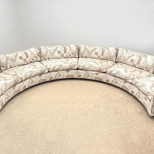 May include: A large, curved sectional sofa upholstered in a cream-colored fabric featuring a repeating geometric pattern in shades of tan and beige. The sofa consists of multiple modular segments with low backrests and deep, cushioned seating areas.