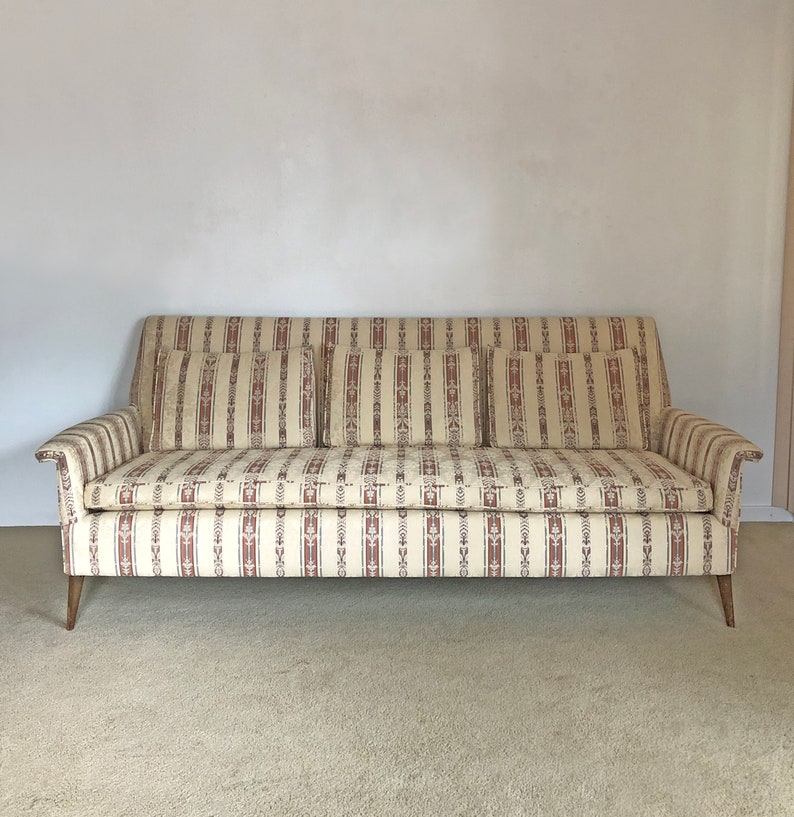 May include: A vintage striped sofa with three pillows. The sofa is upholstered in a beige and brown striped fabric with a floral pattern. The sofa has wooden legs.