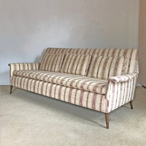 May include: A striped fabric sofa with four pillows. The sofa has a light beige background with thin red stripes and a floral pattern. The sofa has wooden legs.