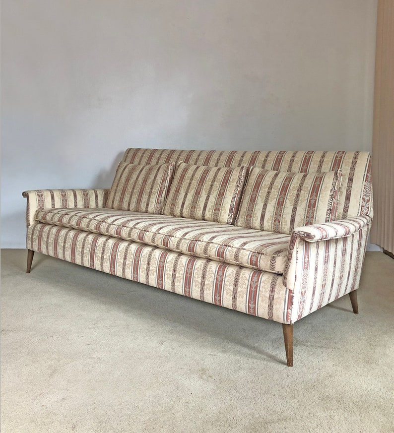 May include: A striped fabric sofa with four pillows. The sofa has a light beige background with thin red stripes and a floral pattern. The sofa has wooden legs.