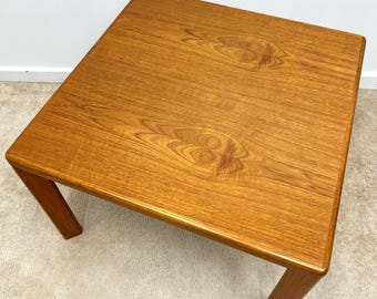 May include: A square, wooden coffee table with a light brown, natural wood finish. The table has four legs and a smooth, flat top.