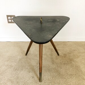 May include: A triangular side table with a dark gray top and three wooden legs with gold accents. The table has a brass ring in the center of the top.