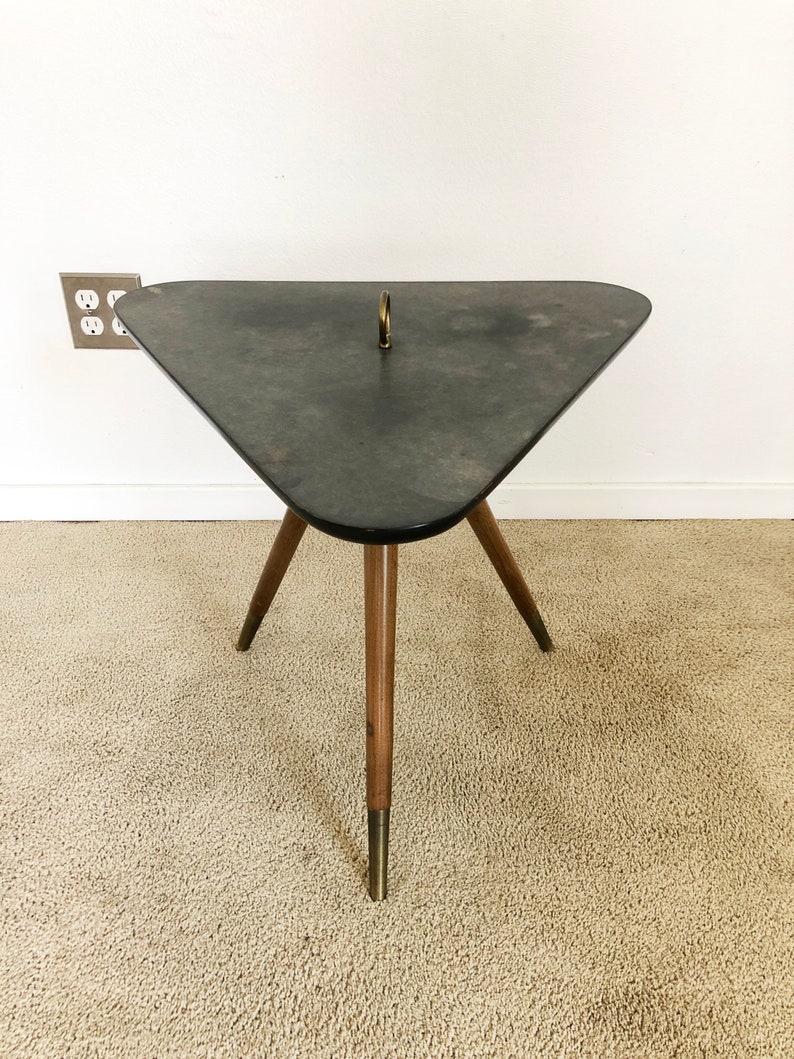 May include: A triangular side table with a dark gray top and three wooden legs with gold accents. The table has a brass ring in the center of the top.