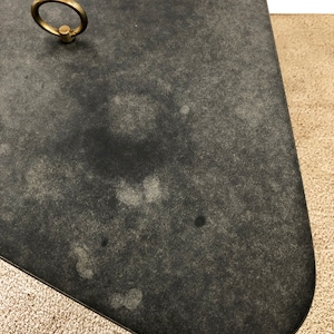 May include: A black, oval-shaped table top with a brass ring handle. The table top has a distressed, aged look with a subtle speckled pattern.