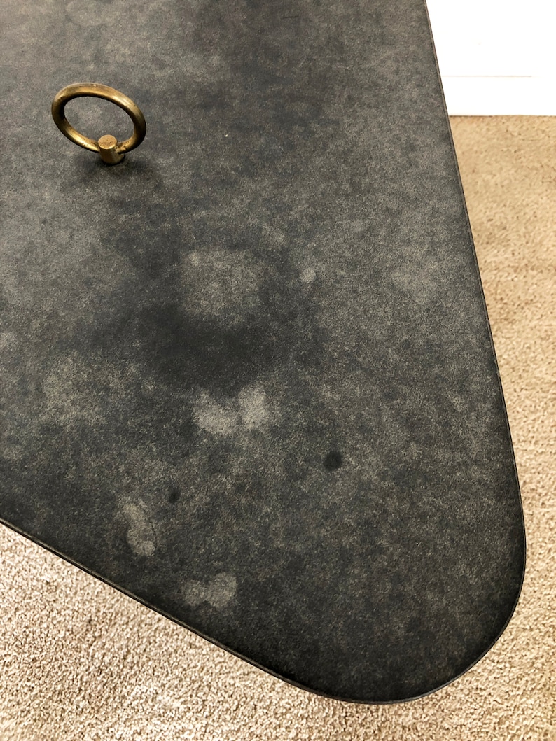 May include: A black, oval-shaped table top with a brass ring handle. The table top has a distressed, aged look with a subtle speckled pattern.