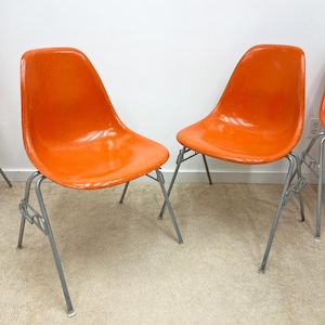 May include: Two orange fiberglass chairs with silver metal legs. The chairs are on a light beige carpet.