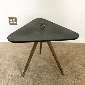 May include: A triangular side table with a dark gray top and a brass ring handle. The table has three wooden legs with brass tips.