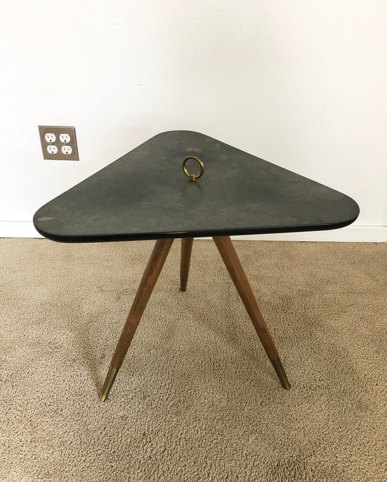 May include: A triangular side table with a dark gray top and a brass ring handle. The table has three wooden legs with brass tips.