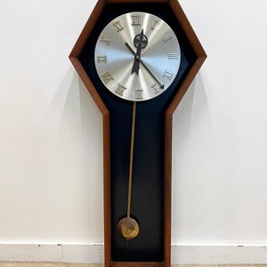 May include: A mid-century modern wall clock with a hexagonal wooden frame. The clock face is silver with black hands and Roman numerals. A long pendulum with a brass-colored weight hangs below. The clock is approximately 30 inches tall.