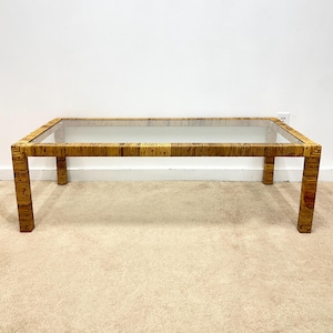 May include: Rectangular coffee table with a clear glass top and a woven rattan frame. The table has a natural, light brown color and four sturdy legs. A stylish piece for a living room or lounge area.