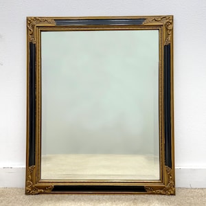 May include: A rectangular mirror with a gold and black ornate frame. The frame has decorative corner accents and a black center section. The mirror is displayed against a white wall and a light-colored carpet.