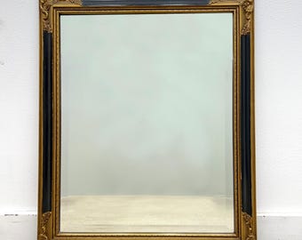May include: A rectangular mirror with a gold and black ornate frame. The frame has decorative corner accents and a black center section. The mirror is displayed against a white wall and a light-coloured carpet.