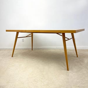 May include: A mid-century modern dining table with a light wood finish. The rectangular tabletop is supported by four angled, tapered legs connected by a stretcher. The table is approximately 72 inches long.