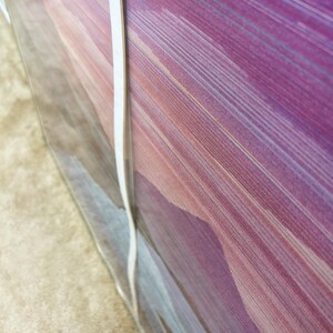 May include: Abstract art print featuring a blend of purple, pink, and gold hues. The artwork showcases a series of diagonal lines, creating a sense of movement and depth. The print is encased in clear plastic, with a white strip along the edge.