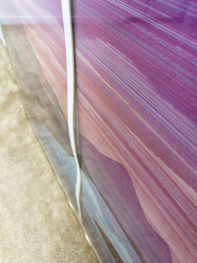 May include: Abstract art print featuring a blend of purple, pink, and gold hues. The artwork showcases a series of diagonal lines, creating a sense of movement and depth. The print is encased in clear plastic, with a white strip along the edge.