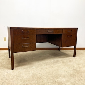 May include: A large, dark wood desk with six drawers and a brown carpet underneath. The desk has a simple design with brass pulls on the drawers.