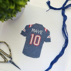 May include: Cross-stitch design of a blue and white football jersey with the name "MAYE" and the number "10" in red. The jersey is on white fabric, with blue thread and scissors nearby. A small green plant is in the background.