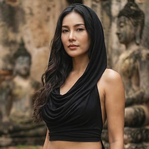 Może przedstawiać: Kobieta w czarnym topie z kapturem i pasujących czarnych spodniach. Top ma dekolt typu cowl i ramiączka typu halter. Tło przedstawia rozmyte kamienne posągi i ceglaną ścianę.