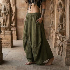 Puede incluir: Una mujer con un top corto negro con cordones y pantalones harén verde oliva. Tiene rastas y tatuajes. Está en un corredor de piedra con pilares y estatuas talladas.